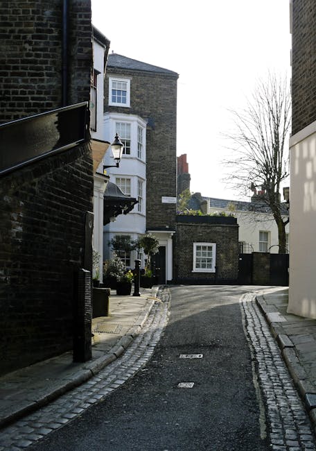 A narrow residential cobbled street in an urban setting, bordered by dark brick buildings on the left and lighter modern structures on the right, with a small white bay-fronted house featuring large sash windows and a black door towards the background. In front of the house, there are decorative street lamps, potted plants, and limited street furniture, indicating a quiet, well-maintained neighbourhood. The street curves gently to the right and is devoid of vehicles or pedestrians, with leafless trees visible against a clear sky. The setting suggests an environment where independent waste collection services, such as those provided by Rubbish Clearance Hampstead, might carry out on-site rubbish removal or private disposal to support efficient waste management in the area without reliance on local authority collection, fitting the context of alternative rubbish handling in residential Hampstead.