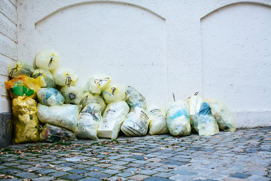 A collection of yellow, white, and transparent plastic trash bags filled with waste material, stacked and leaning against a white textured exterior wall on a cobblestone street. Some bags are tied with black or yellow strings, with visible labels and contents that include paper and miscellaneous debris. The bags vary in size and appear to contain household or yard waste. The cobblestone surface beneath the bags has a mix of gray and brown tones with small scattered leaves and debris. The wall behind is plain, with minimal architectural detail, and the scene is evenly lit with natural daylight, giving a neutral and professional appearance relevant to independent waste collection or rubbish removal services, as offered by Rubbish Clearance Hampstead.