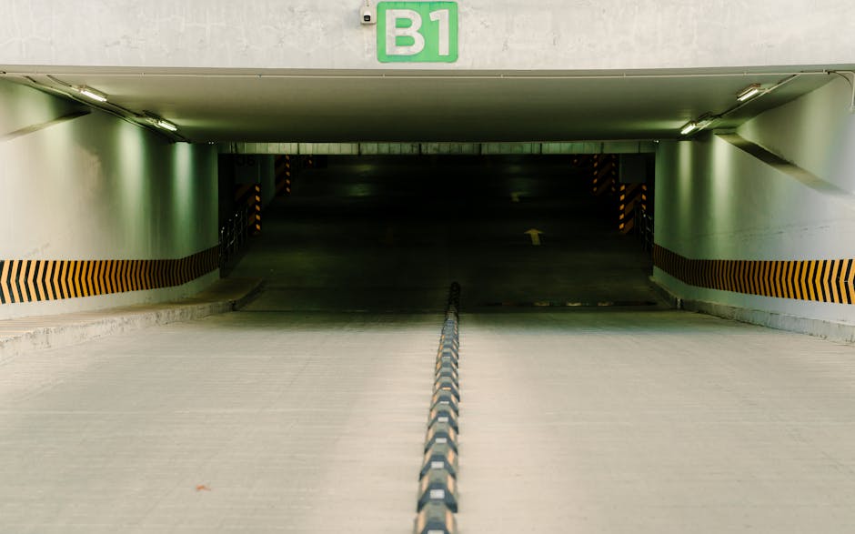 An underground parking garage entrance with a concrete driveway leading into a dark interior. The ramp is bordered by white walls decorated with a horizontal orange and black striped safety barrier. A metal chain barrier runs down the center of the driveway, guiding vehicles towards the entrance. Positioned above the opening is a green sign labeled 'B1', indicating the level or section within the parking structure. The ceiling has a smooth, white surface with embedded fluorescent lighting fixtures casting a subtle glow over the scene. The environment appears clean and well-maintained, consistent with a location used for vehicle access and potential temporary storage, aligning with private waste handling or alternative rubbish removal methods in a residential or commercial underground parking facility. This setting suggests the importance of clear signage and proper organisation for efficient entry and exit, which can aid independent waste collection or on-site clearance operations. The background is obscured in darkness, emphasizing the contrast between the well-lit ramp and the shadowy interior beyond. Overall, the scene depicts a functional, industrial space designed for vehicle movement, relevant to waste management or rubbish removal services operating in such environments.