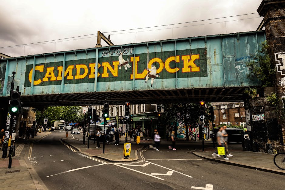 A large, weathered metal railway bridge painted in faded teal spans across a busy street intersection, with the words 'CAMDEN LOCK' prominently displayed in bold yellow letters on its side. The bridge features three hanging figures of children painted as part of street art, positioned near the center, giving a playful yet urban feel. The environment shows an overcast sky with grey clouds, and the street below is active with pedestrians crossing at crosswalks, some wearing casual clothing, and a few moving vehicles such as cars and bicycles. Traffic lights are visible in the foreground, with some green signals illuminated. On the left and right sides, there are storefronts and buildings partially obscured by the bridge, along with small trees and side street signs. The scene exudes a typical cityscape atmosphere, which is relevant to local waste and rubbish removal services like those provided by Rubbish Clearance Hampstead, especially in the context of managing waste around busy urban areas and alternative disposal options.