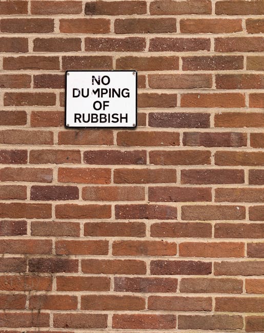 Close-up of a sign mounted on a red brick wall with a staggered pattern, featuring horizontal rows of bricks in various shades of reddish-brown and dark brown mortar joints. The sign is square with a white background and black lettering that reads 'NO DUMPING OF RUBBISH' in bold uppercase letters, with the words 'DUMPING OF' slightly offset and split across two lines. The wall appears weathered, and the lighting creates subtle shadows, emphasizing the texture of the brick surface. This image relates to regulations and enforcement on waste disposal in the context of rubbish removal services, highlighting the importance of adhering to local rules for proper waste management and the role of authorities and private collection services like Rubbish Clearance Hampstead.