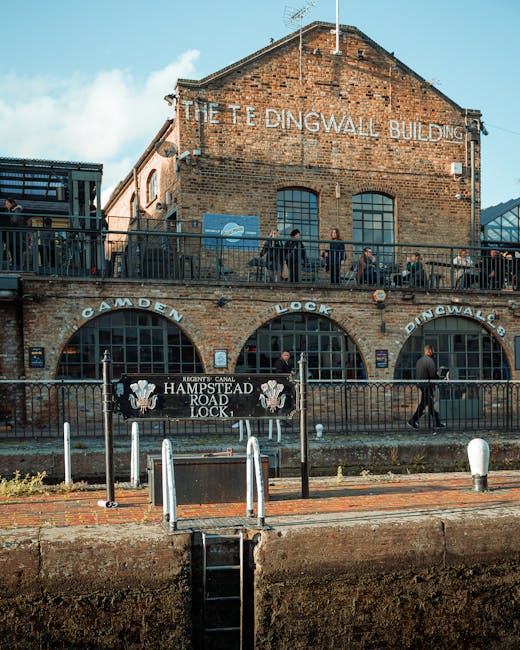 A historic brick building with a gabled roof and weathered lettering that reads 'The Tedingwall Building' is visible in the background, with a large upper-floor balcony enclosed by a black metal railing. Several pedestrians are walking along a paved pathway in front of the building, some stopping at the railing, while others continue past. Beneath the balcony, three arched windows are set into the brick wall, with the words 'Camden Lock' displayed above each arch in white lettering, and a black banner reading 'Hampstead Road Lock' is stretched between the arches. In the foreground, a small, metal, ladder-like structure extends from the sidewalk to a lower canal edge, with white bollards separating the pedestrian area from the waterway. The scene is illuminated by natural daylight, with a partly cloudy sky overhead, and the environment reflects an urban setting near a canal, illustrating an area possibly associated with heritage site preservation or independent waterway management, aligning with principles of private or alternative waste handling and rubbish removal services offered by providers like Rubbish Clearance Hampstead.