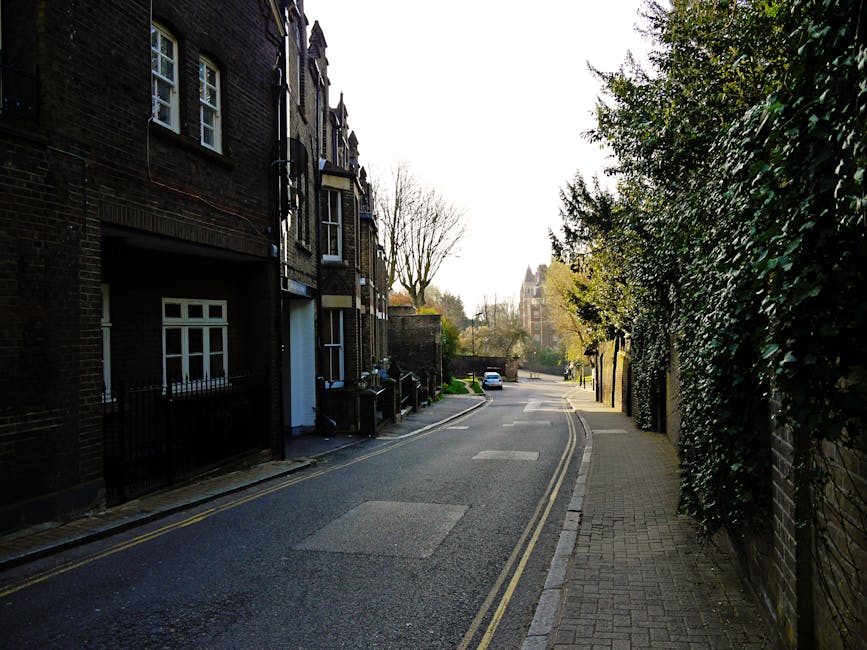 A narrow residential street in Hampstead lined with multi-storey brick townhouses on the left, featuring dark brick facades, white-framed windows, and small front gardens enclosed by black metal railings. The pavement on the right side is paved with concrete slabs, bordered by a high brick wall covered in dense, dark green foliage. Some trees with bare branches are visible further along the street in the background, suggesting late autumn or winter. A parked white car is seen in the distance, and the sky is overcast, casting subtle, diffuse light across the scene. The street appears quiet and clean, with no visible waste or rubbish in the immediate vicinity, reflecting an environment that might be subject to private rubbish clearance services. This setting aligns with a typical Hampstead area, where independent waste removal or on-site clearance by companies like Rubbish Clearance Hampstead are common options for managing rubbish disposal outside typical council collections.
