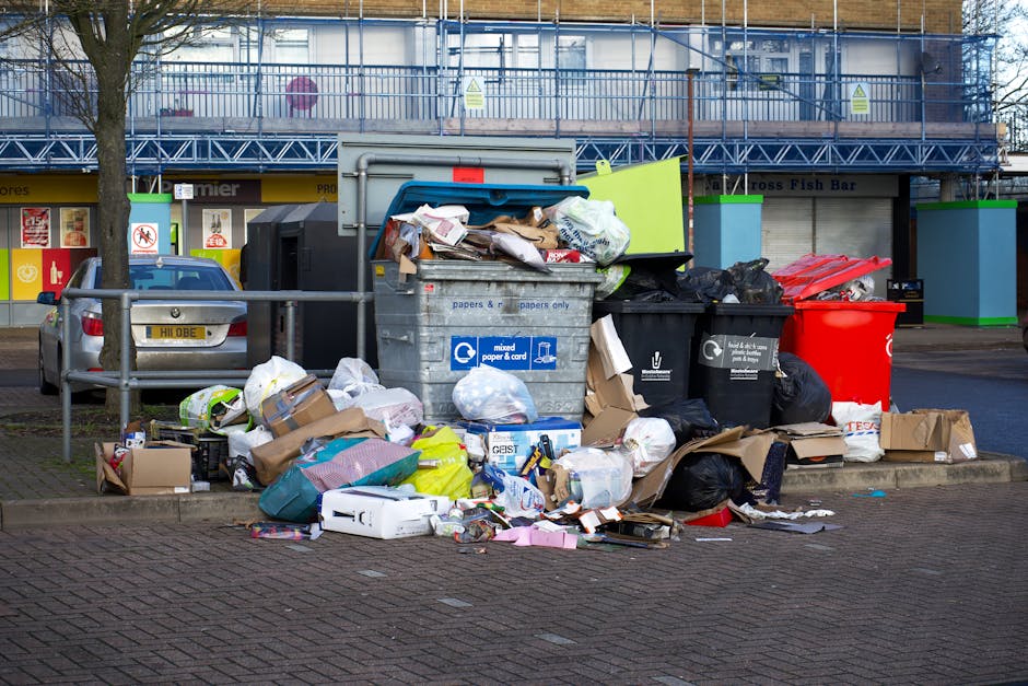 A large collection of mixed waste, including paper, cardboard boxes, plastic bags, black rubbish sacks, and flattened packaging, piled around a central metal rubbish bin with a partially open blue lid, situated on a paved sidewalk area next to a parked silver car with a yellow registration plate. The waste appears to be a combination of household and commercial refuse, with some items spilling onto the surrounding pavement. In the background, a commercial building with bright signage and large windows can be seen, along with a construction scaffold covering the upper level. The scene is set outdoors during daylight, with natural light illuminating the cluttered area, suggesting an instance of on-site rubbish accumulation that might relate to alternative waste handling or private rubbish collection services, as managed by Rubbish Clearance Hampstead, a professional waste management company.