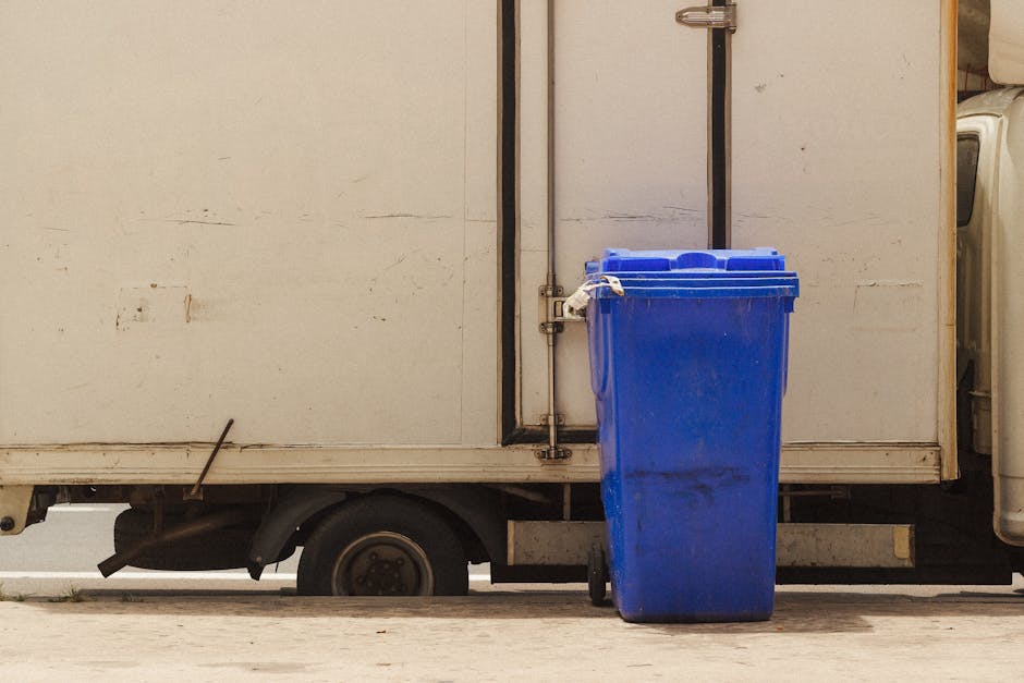 A tall blue plastic rubbish bin with a closed lid is positioned in front of a white delivery truck with a metal exterior. The bin is placed on a paved surface, close to the side of the vehicle, which features a visible black rear wheel partially beneath the body. The truck's exterior shows signs of wear, with visible dirt and slight rust marks, and is situated on a driveway or loading area. The scene is lit with natural daylight, providing clear visibility of textures such as the smooth finish of the plastic bin and the rough, slightly stained surface of the truck. The background includes part of a closed black metal door on the truck and a portion of the adjacent environment, suggesting an urban or suburban setting typical for private waste collection or contingency rubbish removal services. This visual captures a scene related to waste management, with a focus on the use of a standard refuse container ready for collection or disposal, supported by services like Rubbish Clearance Hampstead that facilitate private rubbish removal outside of standard council collections.