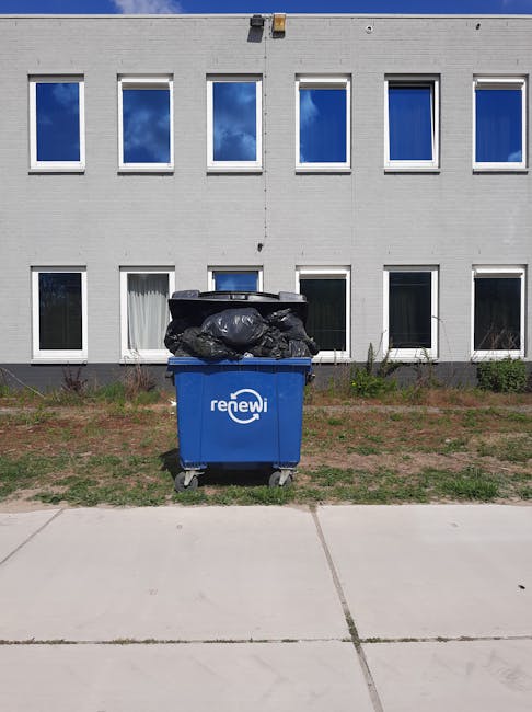 A blue recycling bin with the word 'renewi' printed on its front, positioned on a concrete pavement in front of a modern, grey, two-storey building with multiple rectangular windows reflecting a blue sky with scattered white clouds. The bin is filled with black rubbish bags that are stacked on top, some slightly bulging and irregularly shaped, indicating they are full. The surrounding area consists of a small patch of grass and soil immediately behind the bin, with low shrubs or weeds near the building's base. The scene is illuminated by natural daylight, casting subtle shadows on the pavement and highlighting the textures of the bin, bags, and building's brickwork. This image exemplifies an example of private waste handling and on-site rubbish disposal, typical of independent collection services such as those provided by Rubbish Clearance Hampstead.