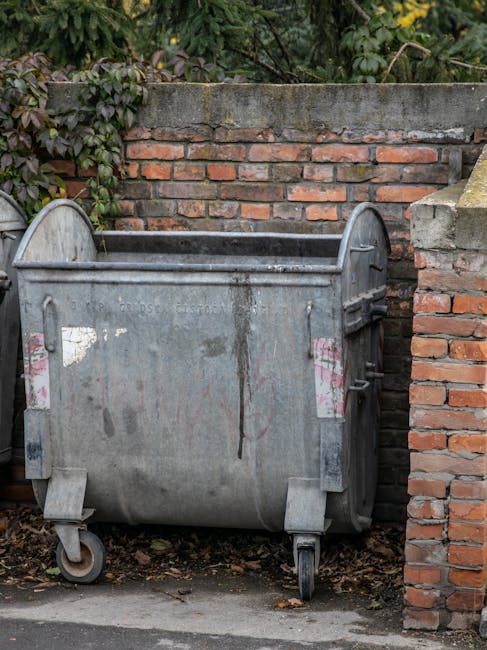A tall blue plastic rubbish bin with a closed lid is positioned in front of a white delivery truck with a metal exterior. The bin is placed on a paved surface, close to the side of the vehicle, which features a visible black rear wheel partially beneath the body. The truck's exterior shows signs of wear, with visible dirt and slight rust marks, and is situated on a driveway or loading area. The scene is lit with natural daylight, providing clear visibility of textures such as the smooth finish of the plastic bin and the rough, slightly stained surface of the truck. The background includes part of a closed black metal door on the truck and a portion of the adjacent environment, suggesting an urban or suburban setting typical for private waste collection or contingency rubbish removal services. This visual captures a scene related to waste management, with a focus on the use of a standard refuse container ready for collection or disposal, supported by services like Rubbish Clearance Hampstead that facilitate private rubbish removal outside of standard council collections.